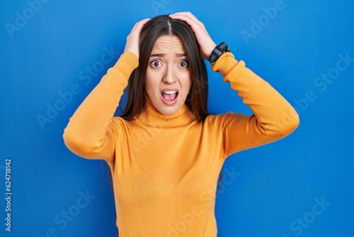 Young brunette woman standing over blue background crazy and scared with hands on head, afraid and surprised of shock with open mouth © Krakenimages.com