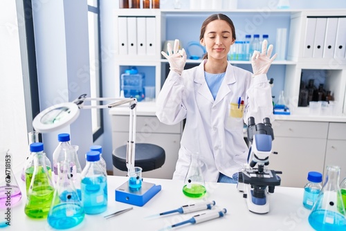Young brunette woman working at scientist laboratory relax and smiling with eyes closed doing meditation gesture with fingers. yoga concept.