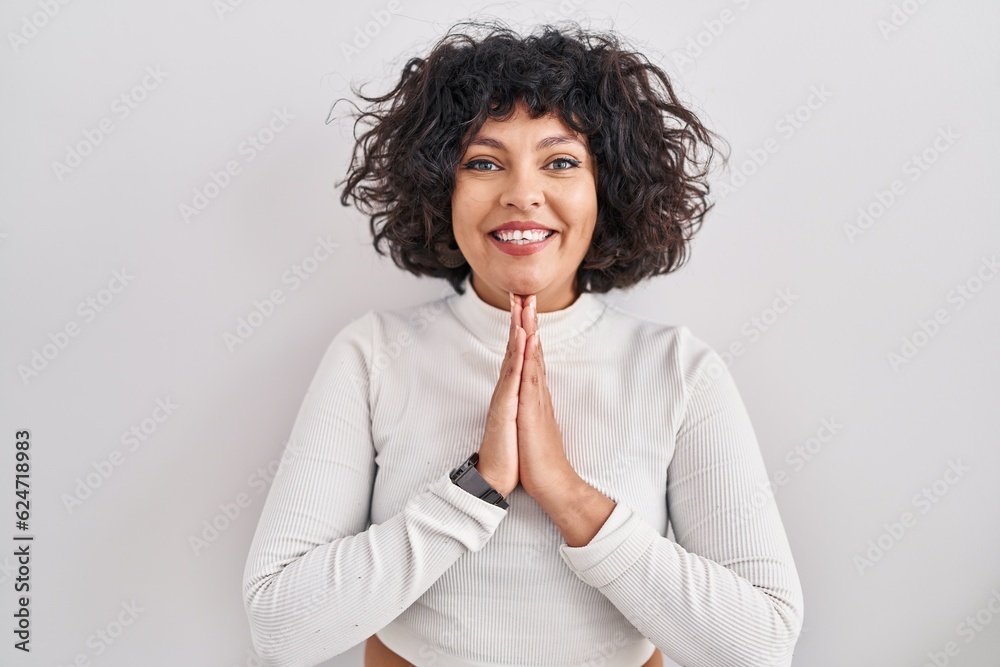 Hispanic woman with curly hair standing over isolated background praying with hands together asking for forgiveness smiling confident. Hispanic woman with curly hair standing over isolated background praying with hands together asking for forgiveness smiling confident.