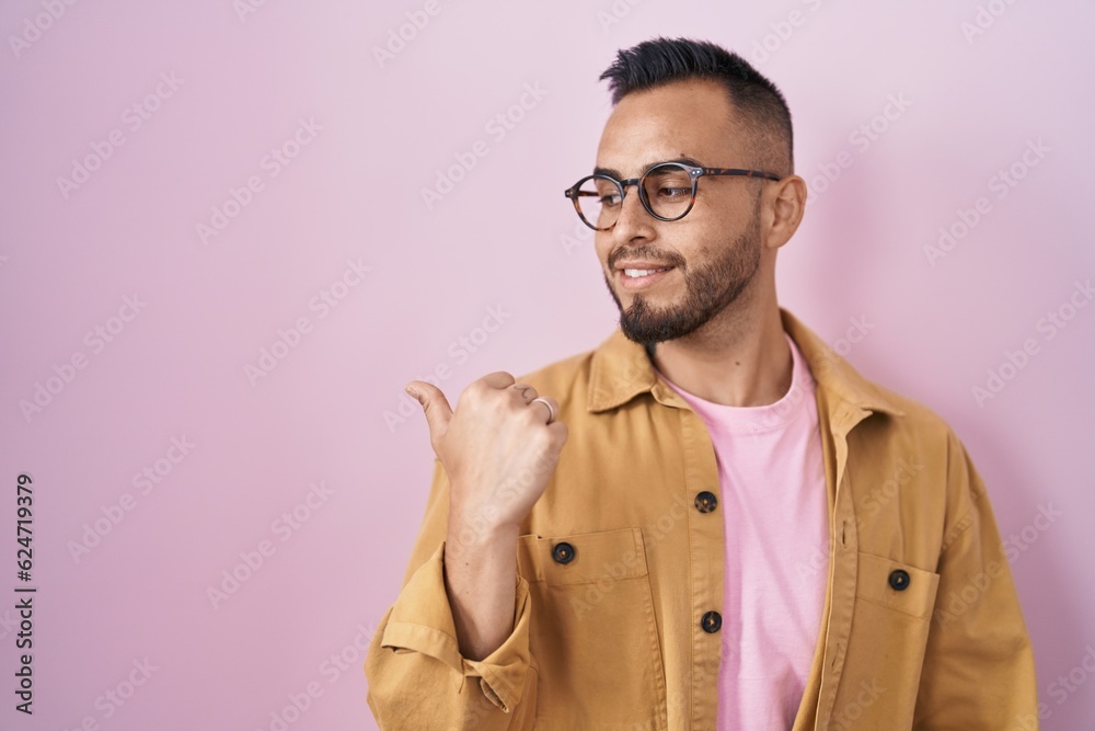 Young hispanic man standing over pink background smiling with happy face looking and pointing to the side with thumb up. Young hispanic man standing over pink background smiling with happy face looking and pointing to the side with thumb up.