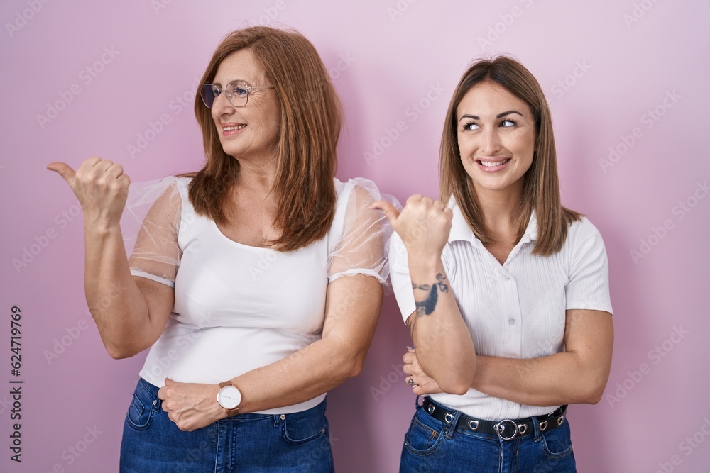 Hispanic mother and daughter wearing casual white t shirt over pink background smiling with happy face looking and pointing to the side with thumb up. Hispanic mother and daughter wearing casual white t shirt over pink background smiling with happy face looking and pointing to the side with thumb up.