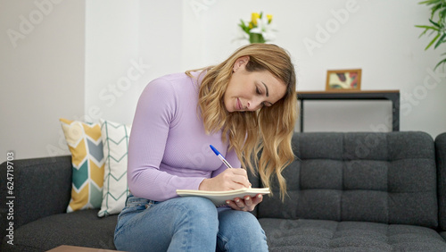 Young beautiful hispanic woman writing on notebook sitting on sofa thinking at home