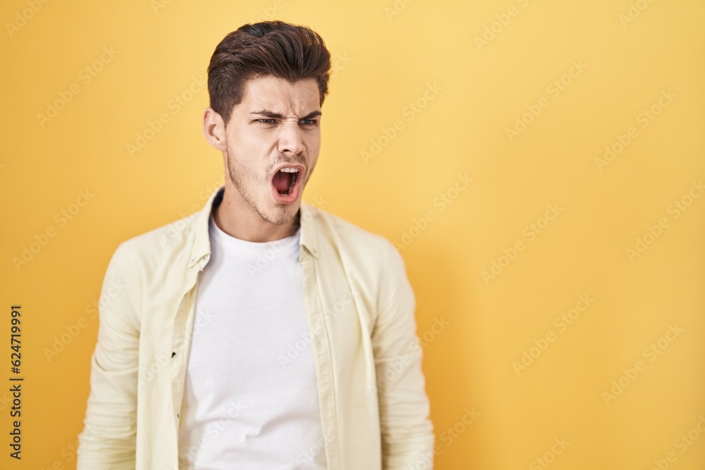 Young hispanic man standing over yellow background angry and mad screaming frustrated and furious, shouting with anger. rage and aggressive concept.