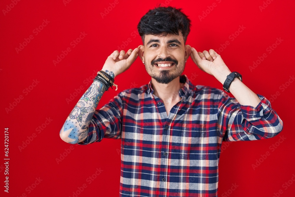 Young hispanic man with beard standing over red background smiling pulling ears with fingers, funny gesture. audition problem