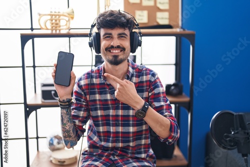 Young hispanic man with beard showing smartphone screen at music studio smiling happy pointing with hand and finger