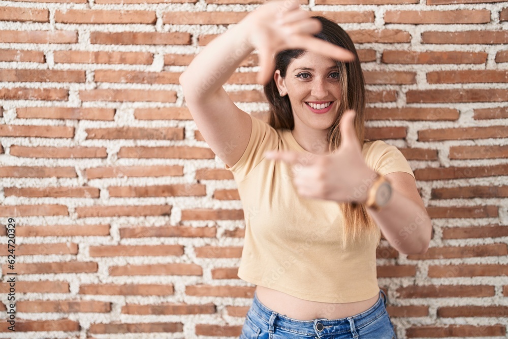 Young brunette woman standing over bricks wall smiling making frame with hands and fingers with happy face. creativity and photography concept. Young brunette woman standing over bricks wall smiling making frame with hands and fingers with happy face. creativity and photography concept.