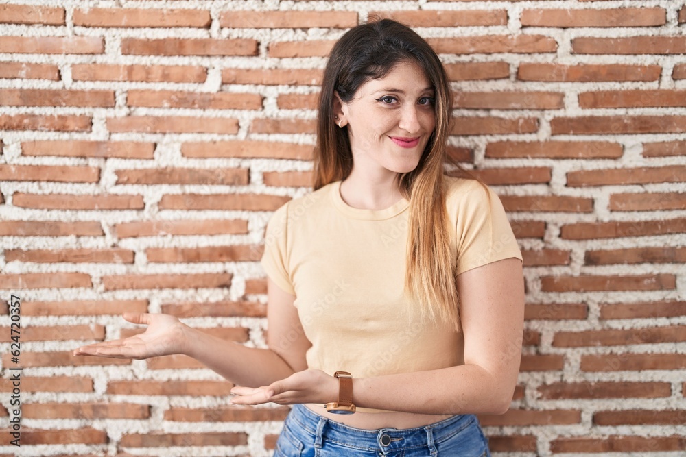 Young brunette woman standing over bricks wall inviting to enter smiling natural with open hand