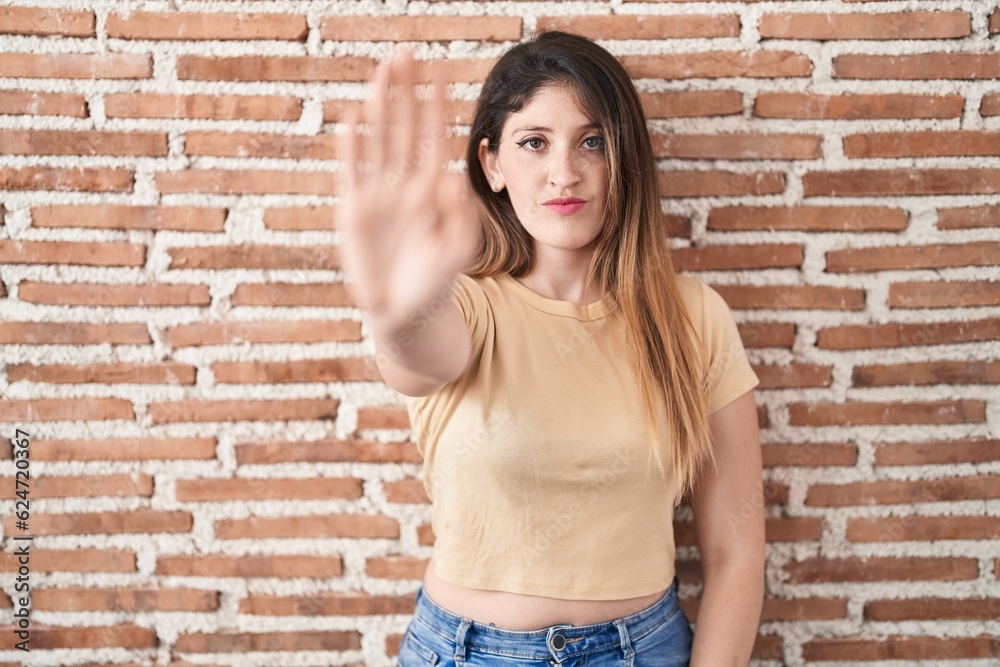 Young brunette woman standing over bricks wall doing stop sing with palm of the hand. warning expression with negative and serious gesture on the face. Young brunette woman standing over bricks wall doing stop sing with palm of the hand. warning expression with negative and serious gesture on the face.