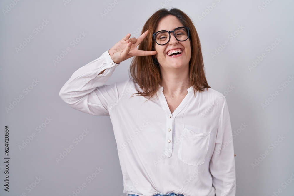 Brunette woman standing over white isolated background doing peace symbol with fingers over face, smiling cheerful showing victory Brunette woman standing over white isolated background doing peace symbol with fingers over face, smiling cheerful showing victory