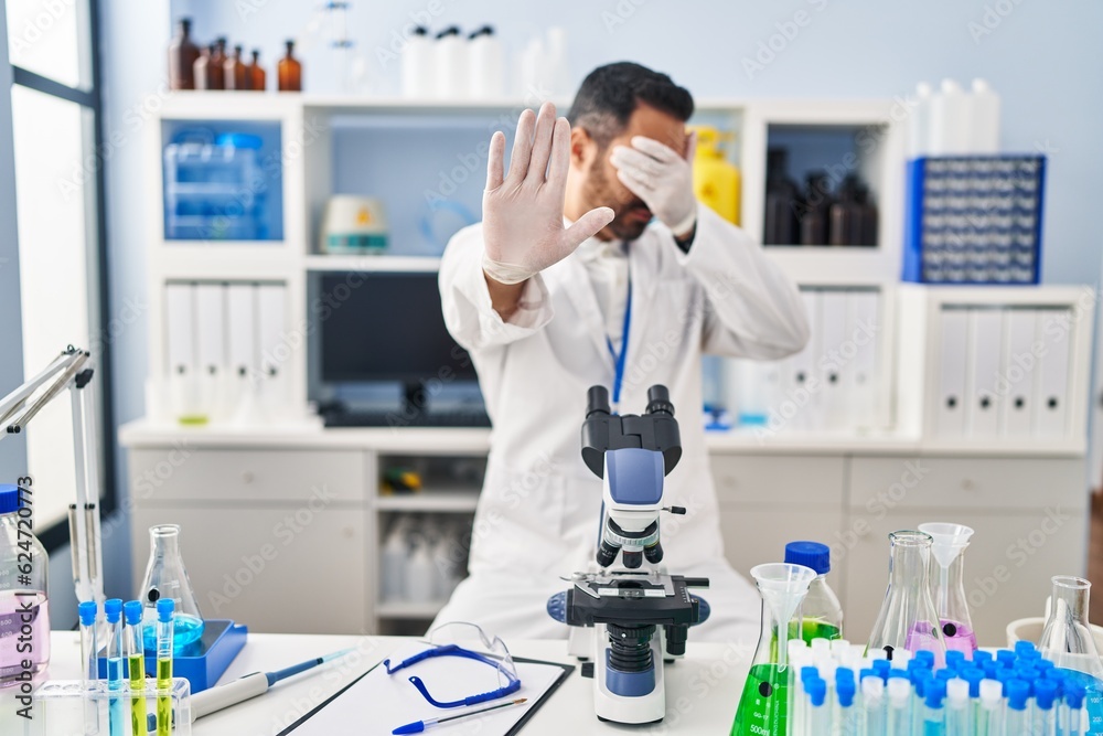 Young hispanic man with beard working at scientist laboratory covering eyes with hands and doing stop gesture with sad and fear expression. embarrassed and negative concept. Young hispanic man with beard working at scientist laboratory covering eyes with hands and doing stop gesture with sad and fear expression. embarrassed and negative concept.
