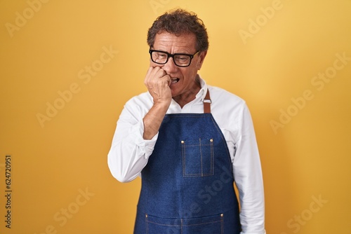 Middle age hispanic man wearing professional cook apron looking stressed and nervous with hands on mouth biting nails. anxiety problem.