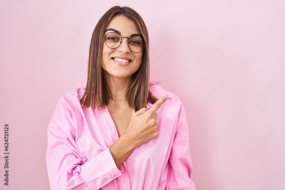 Young hispanic woman wearing glasses standing over pink background cheerful with a smile on face pointing with hand and finger up to the side with happy and natural expression Young hispanic woman wearing glasses standing over pink background cheerful with a smile on face pointing with hand and finger up to the side with happy and natural expression