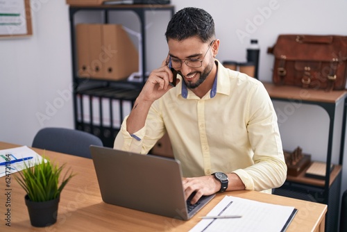 Young arab man business worker using laptop talking on smartphone at office