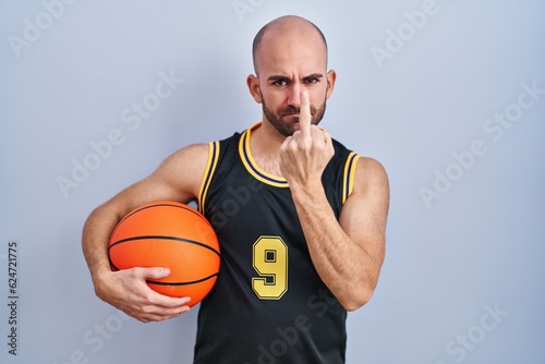 Young bald man with beard wearing basketball uniform holding ball showing middle finger, impolite and rude fuck off expression