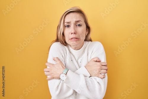 Young caucasian woman wearing white sweater over yellow background shaking and freezing for winter cold with sad and shock expression on face