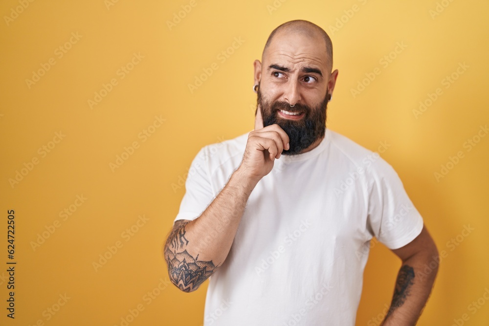 Young hispanic man with beard and tattoos standing over yellow background thinking worried about a question, concerned and nervous with hand on chin