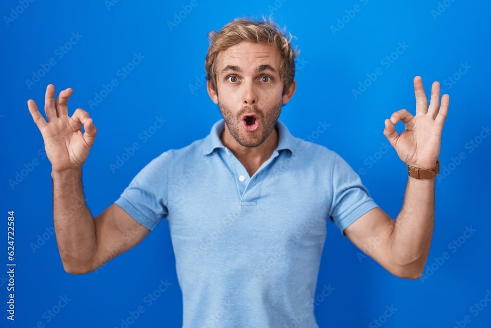Caucasian man standing over blue background looking surprised and shocked doing ok approval symbol with fingers. crazy expression Caucasian man standing over blue background looking surprised and shocked doing ok approval symbol with fingers. crazy expression