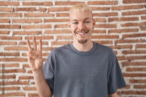 Young caucasian man standing over bricks wall showing and pointing up with fingers number three while smiling confident and happy.