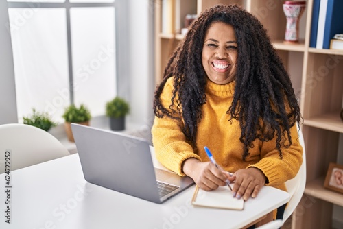 African american woman using laptop writing on notebook at home