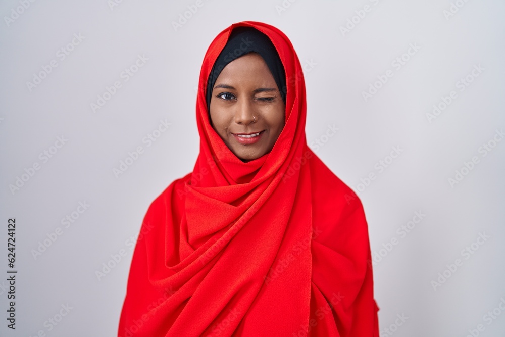 Young arab woman wearing traditional islamic hijab scarf winking looking at the camera with sexy expression, cheerful and happy face. Young arab woman wearing traditional islamic hijab scarf winking looking at the camera with sexy expression, cheerful and happy face.