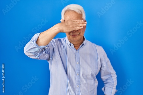 Hispanic senior man wearing glasses covering eyes with hand, looking serious and sad. sightless, hiding and rejection concept
