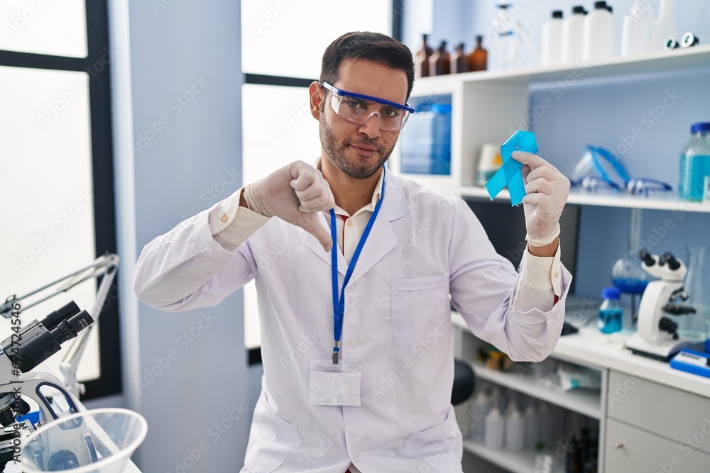 Young hispanic man with beard working at scientist laboratory holding blue ribbon with angry face, negative sign showing dislike with thumbs down, rejection concept Young hispanic man with beard working at scientist laboratory holding blue ribbon with angry face, negative sign showing dislike with thumbs down, rejection concept