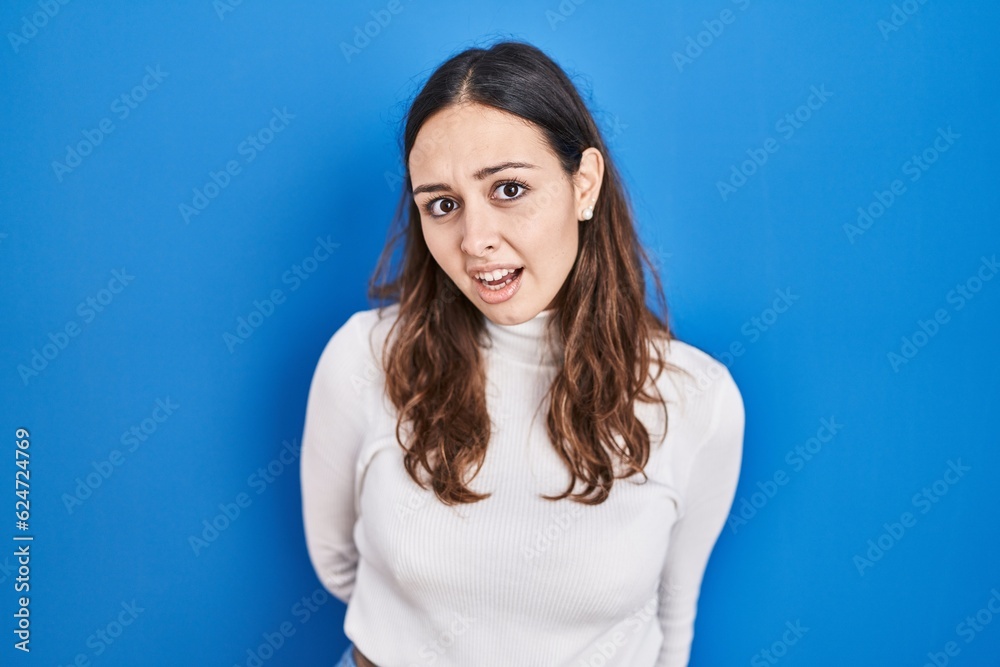 Young hispanic woman standing over blue background in shock face, looking skeptical and sarcastic, surprised with open mouth Young hispanic woman standing over blue background in shock face, looking skeptical and sarcastic, surprised with open mouth