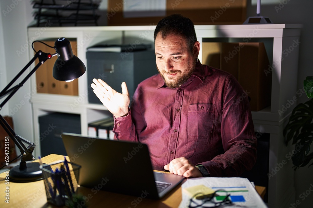 Plus size hispanic man with beard working at the office at night smiling cheerful presenting and pointing with palm of hand looking at the camera. Plus size hispanic man with beard working at the office at night smiling cheerful presenting and pointing with palm of hand looking at the camera.