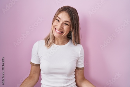 Blonde caucasian woman standing over pink background smiling cheerful with open arms as friendly welcome  positive and confident greetings