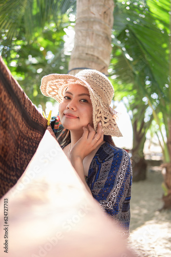 A young Asian woman wearing traditional clothes made of woven cloth and combined with batik cloth while holding a hat against a tree background during the day