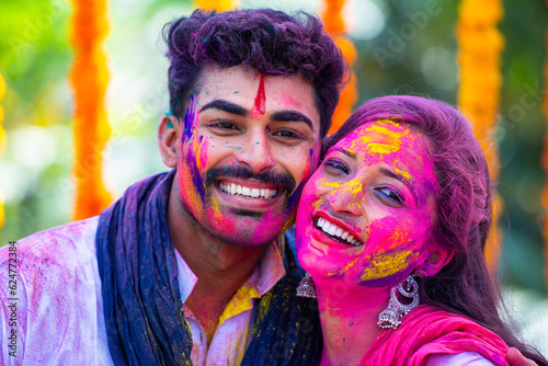 Close up shot of happy young indian couple with holi colour on face by looking at camera during holi festival celebration - concept of freedom  togetherness and indian culture