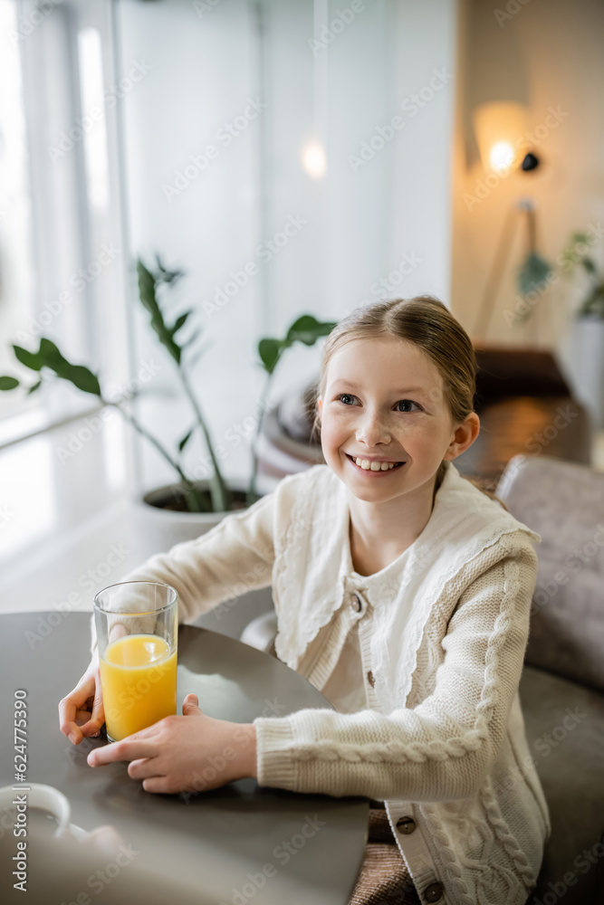 happy preteen girl holding glass of orange juice while sitting in cafe, positivity and joy, round table, smart casual attire, blurred plant on background happy preteen girl holding glass of orange juice while sitting in cafe, positivity and joy, round table, smart casual attire, blurred plant on background