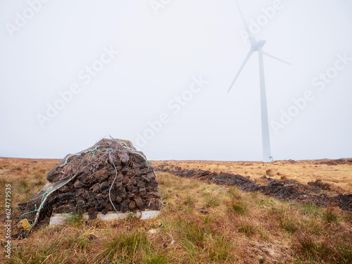 Pile of peat fossil fuel in foreground, wind power turbine in a fog in the background. Old and new source of energy. Ecology concept. Heat producing and energy producing source of energy. photo
