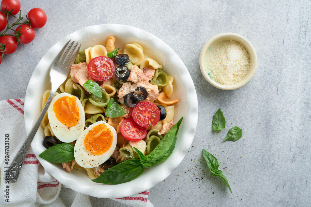 Italian pasta salad. Orecchiette pasta with tuna, tomato cherry, olive, basil and parmesan cheese in plate on grey stone or concrete background. Traditional Italian cuisine. Flat lay. Copy space.