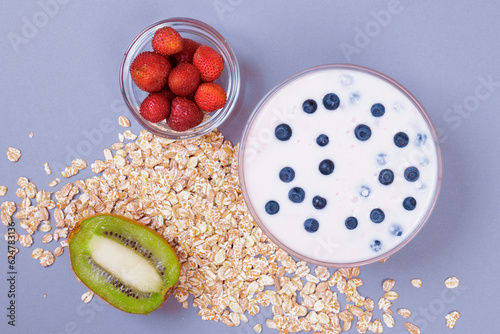 Glass bowl of yogurt with fresh blueberries, bowl with strawberries, half of kiwi and a lot of scattered cereals on gray background. Top view