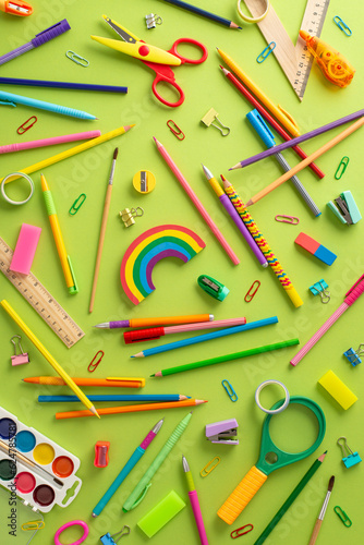 Emphasize the back-to-school theme: Vertical top view of scattered color pencils, watercolor paints, plasticine, scissors, ruler, clips, magnifying glass, erasers and more on pastel green background