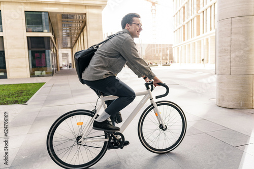 Fototapeta Naklejka Na Ścianę i Meble -  Hipster male freelancer riding to work on a bicycle with a bag, to the office on an eco-transport, millennial in stylish clothes.