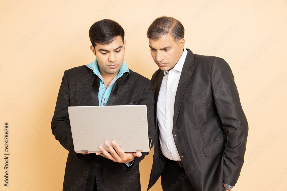 Two indian businessman wearing black formal suit working on laptop isolated over beige background. Business and corporate concept. Two indian businessman wearing black formal suit working on laptop isolated over beige background. Business and corporate concept.