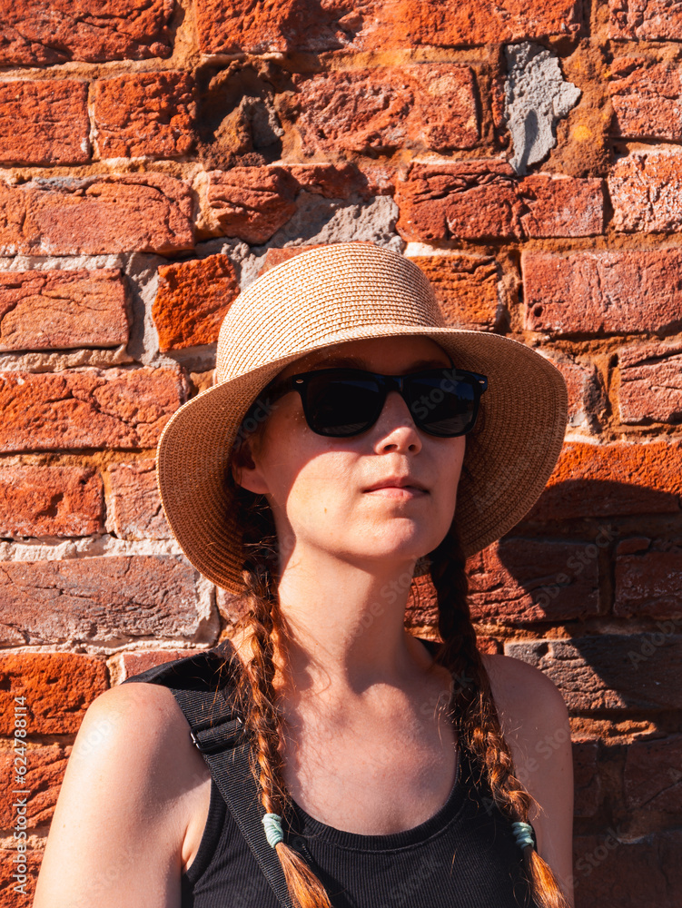 Young woman in straw hat portrait on red brick wall urban background. Millennial girl walking on summer city street with bright sunshine. Stylish confident authentic lady in black sunglasses outdoors.