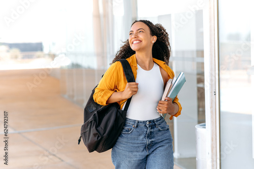 Happy lovely curly haired brazilian or hispanic female student, with a backpack, hold books and notebooks in her hand, walking near the university campus, looks away and smile, finished school day photo