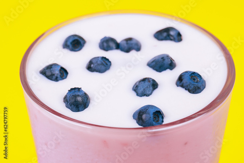 Glass of cold milkshake with fresh blueberries on top. Drink in a glass with a straw on a yellow background.