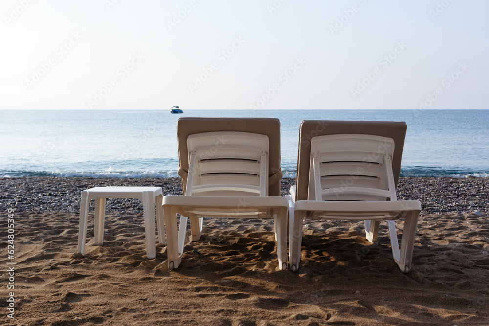 Two sun loungers and a table on a beach. Blue sea background. Silhouette of motorboat on the horizon. Vacatoin concept