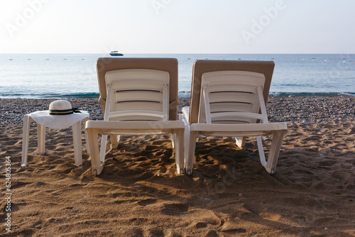 Two sun loungers and a table with a straw hat on a beach. Blue sea background. Silhouette of motorboat on the horizon. Vacatoin  © Irina