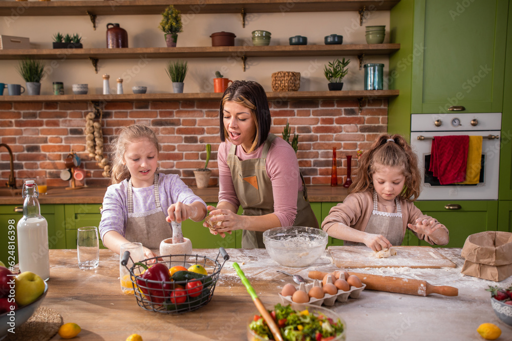 Beautiful woman and her two daughters have a fun time at the kitchen island at home they playing with flour and mix the dough to prepare the delicious biscuits together