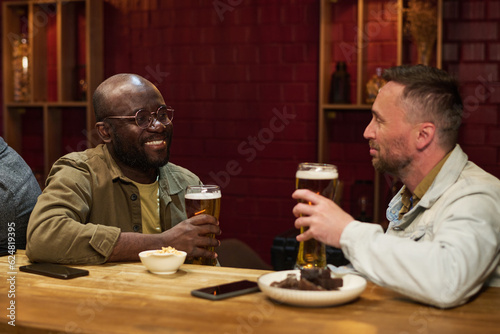 Happy young African American man with glass of beer looking at his buddy during conversation by bar counter while enjoying weekend