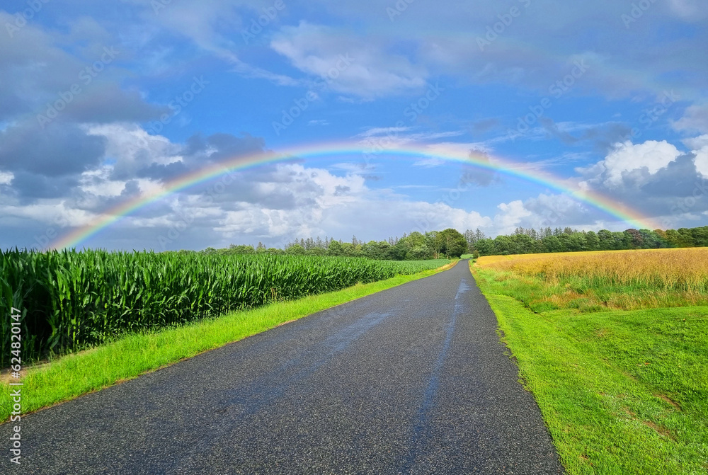 Rainbow in the blue sky hanging over a small road in the Danish countryside with agricultural fields