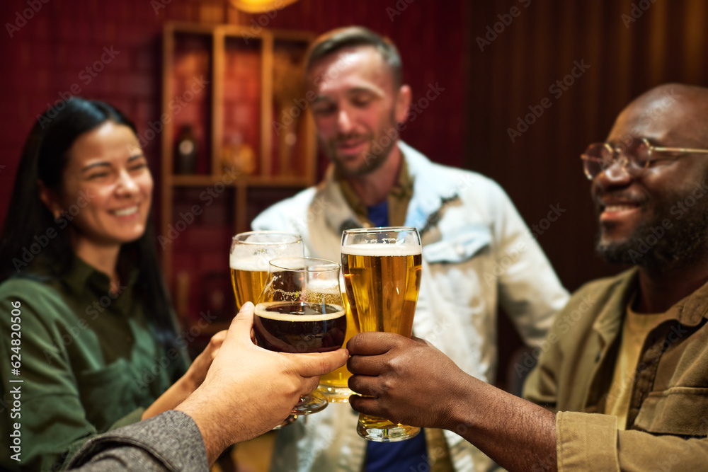 Focus on hands of happy young friends clinking with glasses of beer while standing in front of camera in bar and enjoying party or weekend