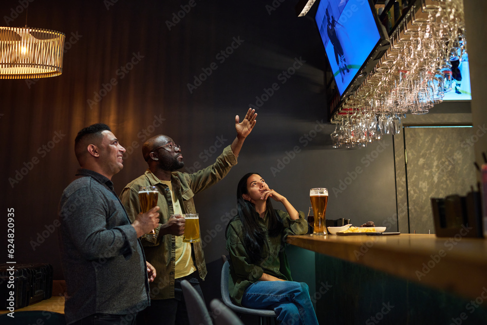 Side view of young intercultural friends having beer and watching hockey broadcast while one of them expressing tension and pointing at screen