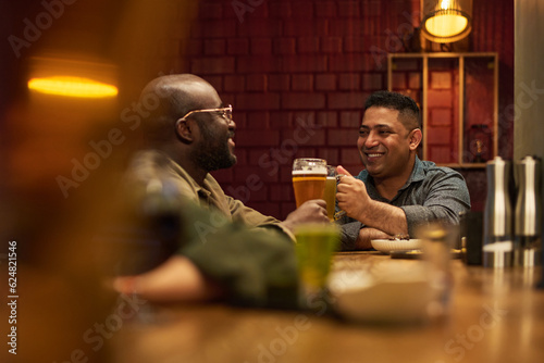 Two happy young intercultural buddies clinking with glasses of foaming beer and looking at one another while sitting by bar counter