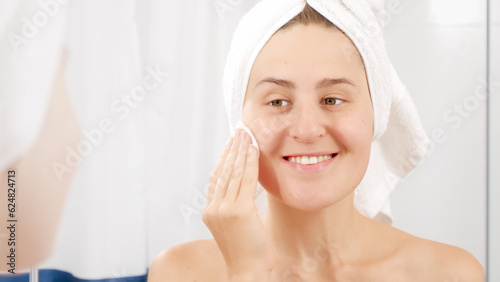 Smiling woman in bath towel cleaning face with lotion after having shower. Concept of beautiful female, makeup at home, skin care and domestic beauty industry.